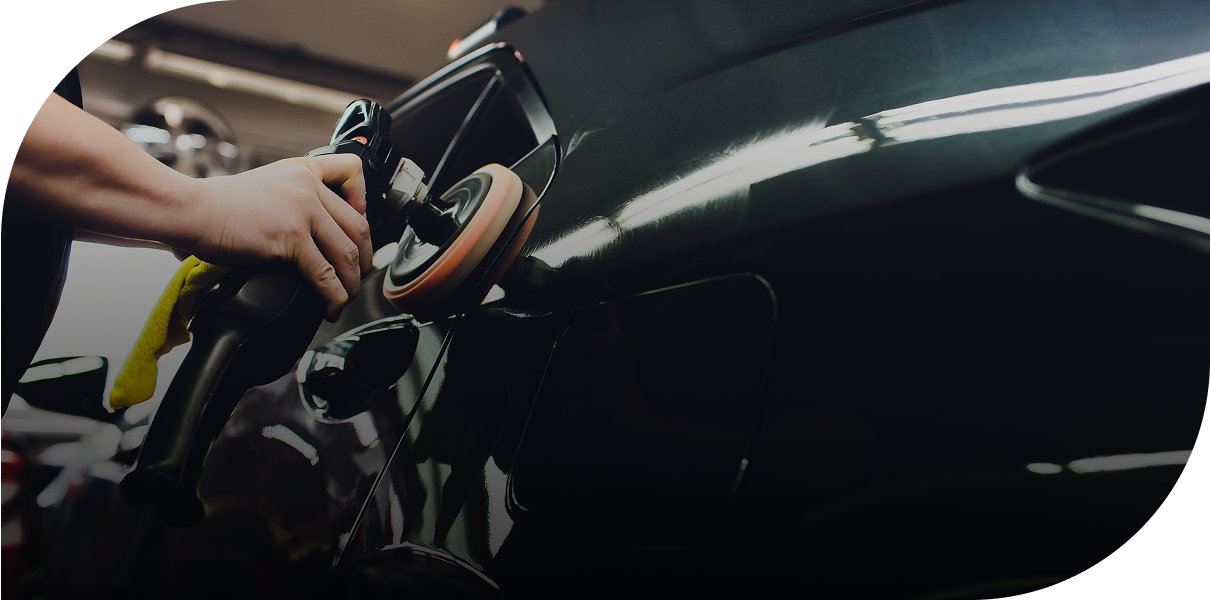 Person polishing a dark-colored car with a buffer in a garage setting.