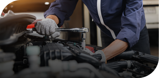 Mechanic wearing gloves and blue coveralls using a wrench to work on a car engine under the hood.