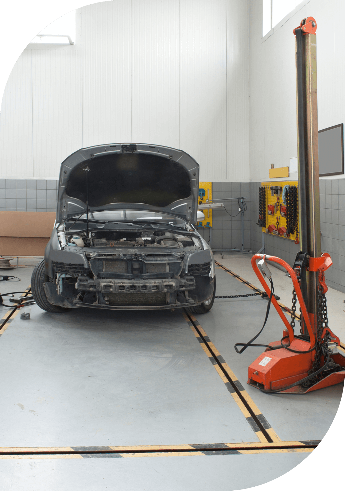 A car with an open hood in an auto repair shop, with a damaged front bumper and a red hydraulic lift on the right.