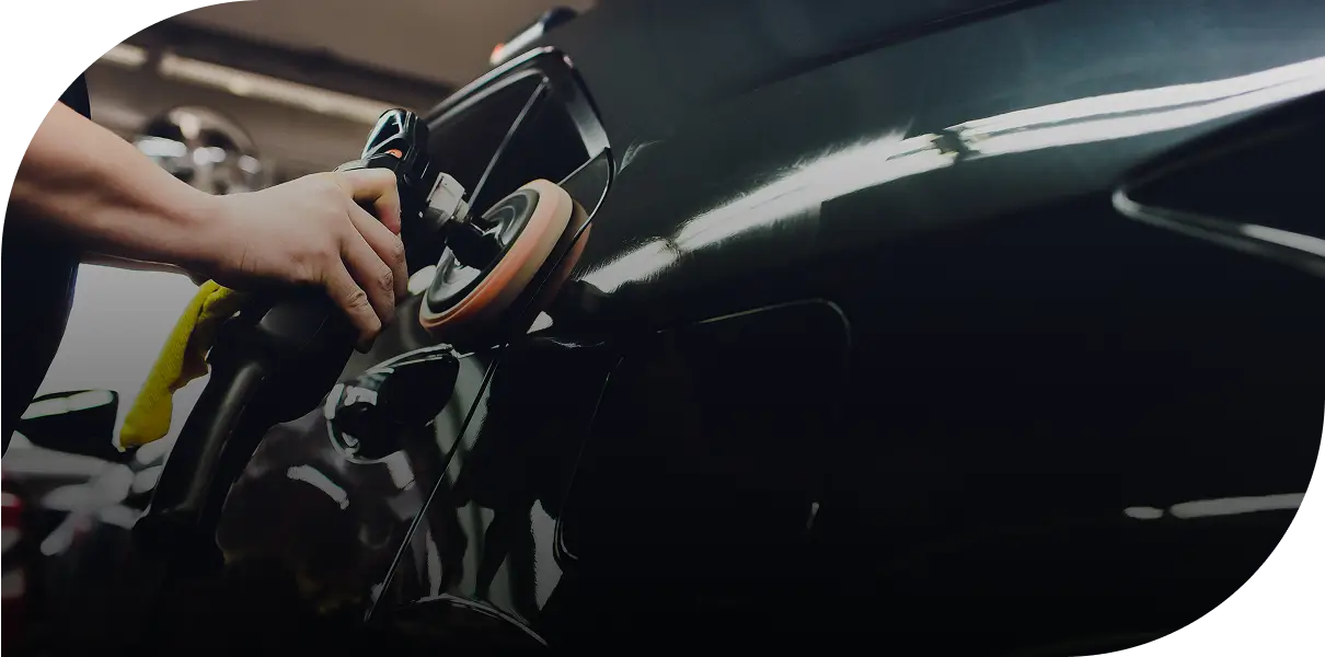 Person polishing a dark-colored car with a buffer in a garage setting.