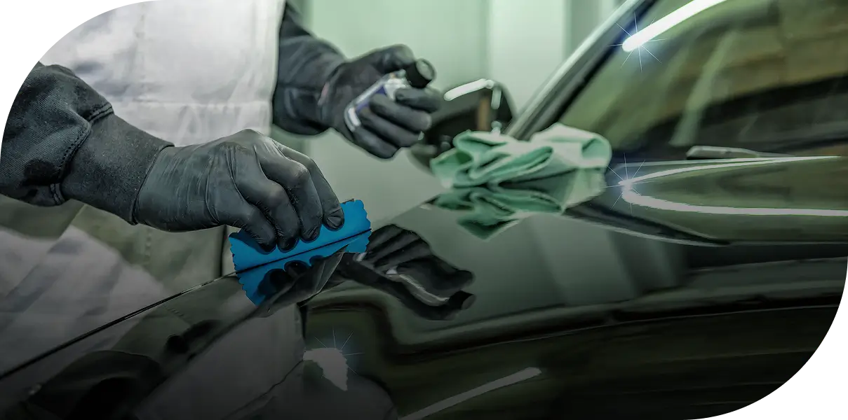 Person wearing gloves applies wax to a black car's polished surface with a blue cloth, while holding a spray bottle.